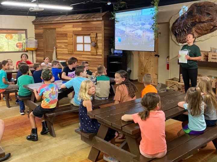 A Science Center educator speaking to a group of children seated at picnic tables, beneath a mounted Tyrannosaurus skull.