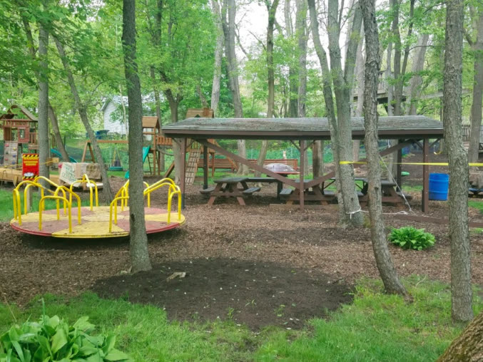 The outdoor play park among the trees, with a merry-go-round, swings, a climbing structure and a covered picnic pavilion.