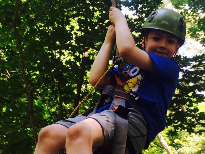 A boy in a helmet and harness riding the zip line through the trees, smiling at the camera.