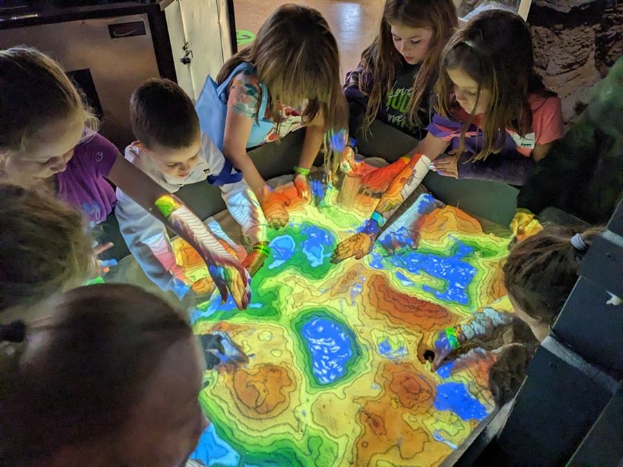 Children gathered around the augmented reality sandbox, hands in the sand as coloured contour lines and blue water are projected onto it.