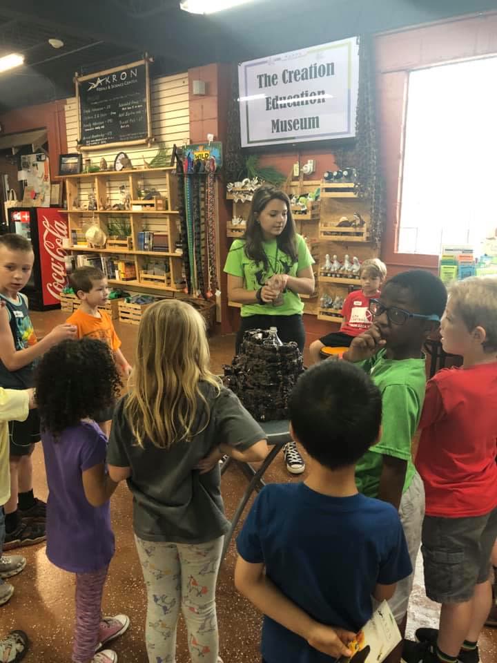 A guide in a green shirt talking with a group of children beneath the Creation Education Museum sign.