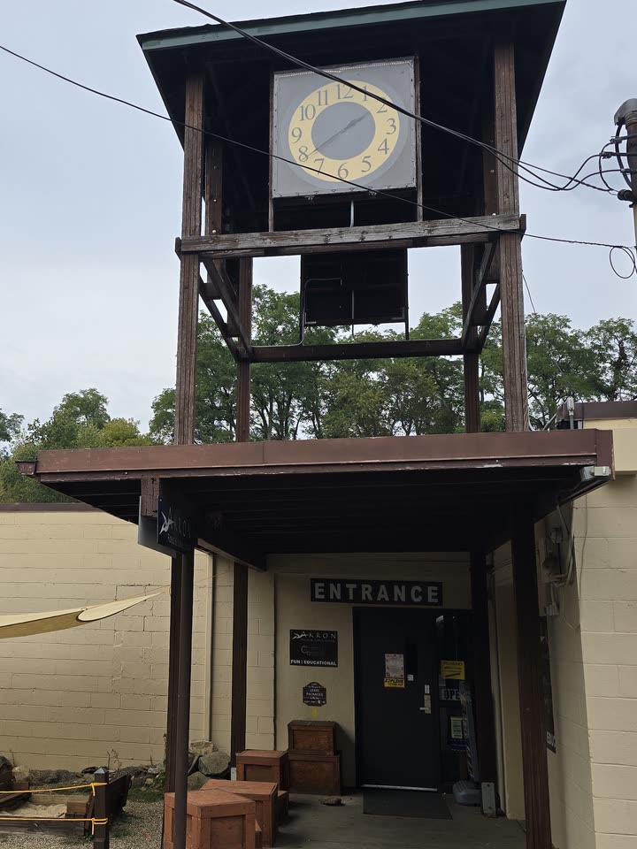 A wooden clock tower standing over a covered walkway, with a doorway beneath it under a sign reading ENTRANCE.