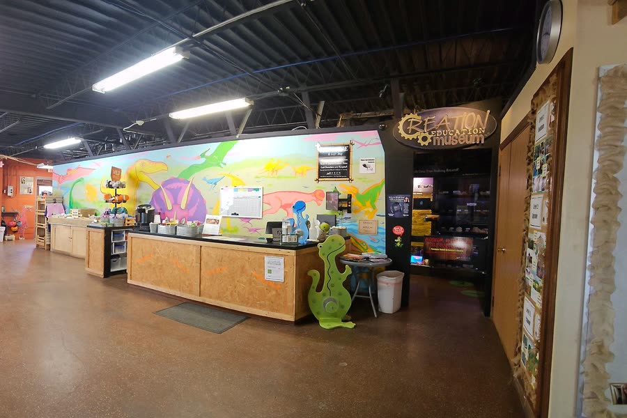 The front counter inside the building, with a painted dinosaur mural behind it and the Creation Education Museum sign over a doorway to one side.